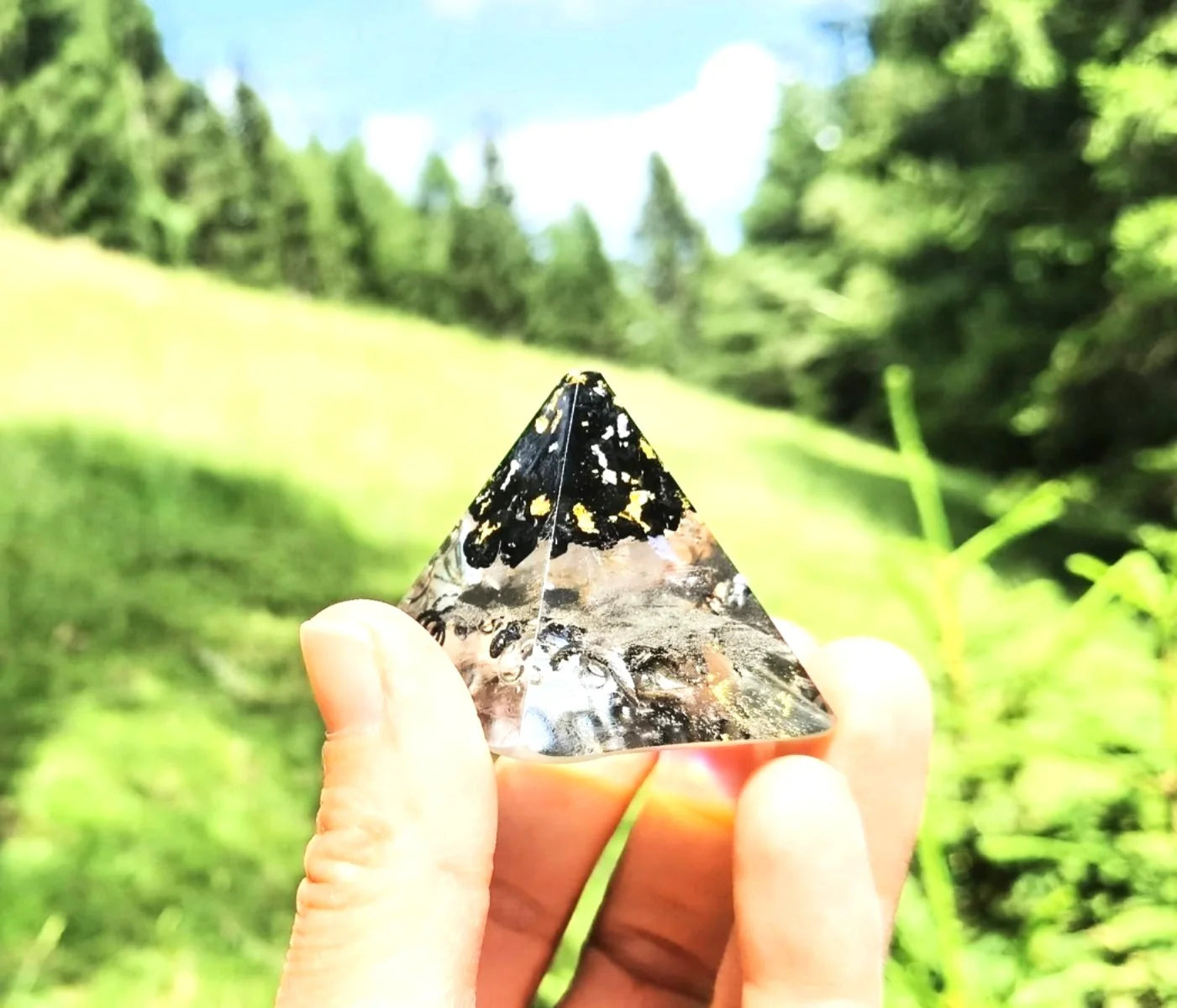 Handgefertigte Mini-Orgonit-Pyramide mit Schungit, Gold und Kristallen, gehalten vor einem grünen Naturhintergrund.