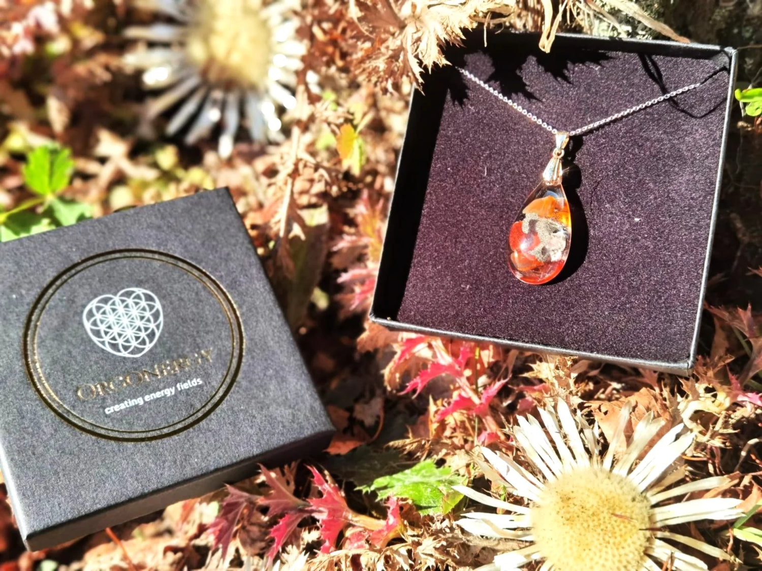 Handgefertigter Orgon-Anhänger in Tropfenform mit leuchtenden Edelsteinen, präsentiert in einer eleganten schwarzen Schmuckbox mit dem Logo von Orgonergy, umgeben von natürlicher Dekoration. Perfekt für Harmonie, Schutz und Energie.