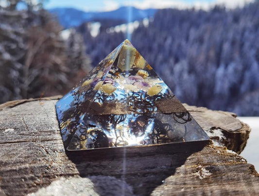 Vorderansicht der Lotusblume Orgon-Pyramide mit Aquamarin, Amethyst und Bergkristall auf einem Holzstamm vor einer verschneiten Berglandschaft.